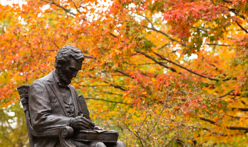 Photograph taken during autumn, showing statue of Lincoln with quill in hand, on Gettysburg campus, fall folliage in background