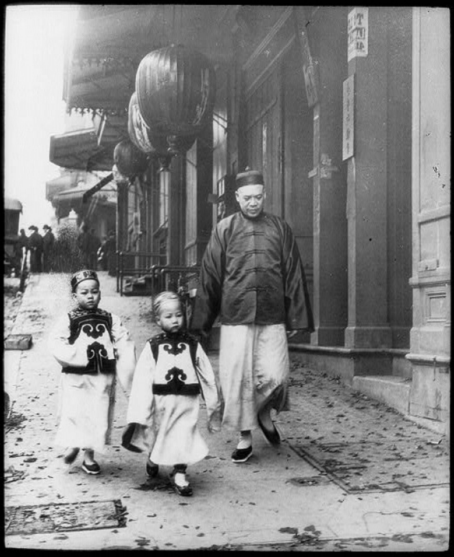 Black and white photo. Adult male walking with two children through San Fransisco's Chinatown in 1896. They are dressed in traditional clothing. (Library of Congress)
