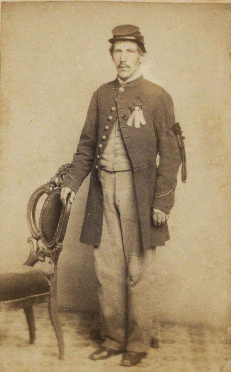 Photograph showing a Civil War veteran wearing a mourning ribbon, a forage hat and his right hand rests on a chair.