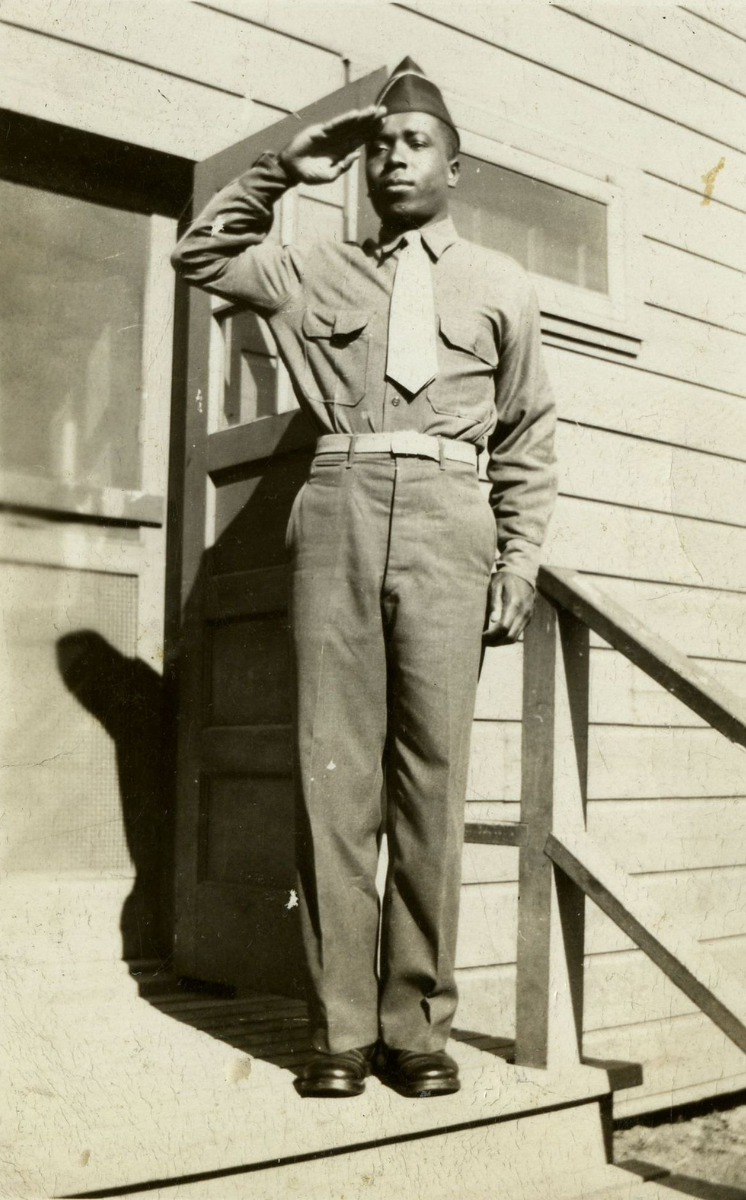Photograph of a World War II African American soldier standing and saluting on a porch, possibly barracks.