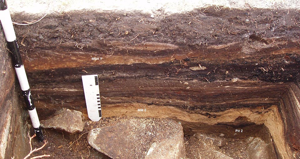 Excavation wall at Cascade Pass shows layers of soil, volcanic ashes, and ancient campfire charcoal spanning 9,000 years (thick black layer that runs across the middle); ground surface at top, glacial boulders at bottom (Photo courtesy of Robert R. Mierendorf, September 2005)