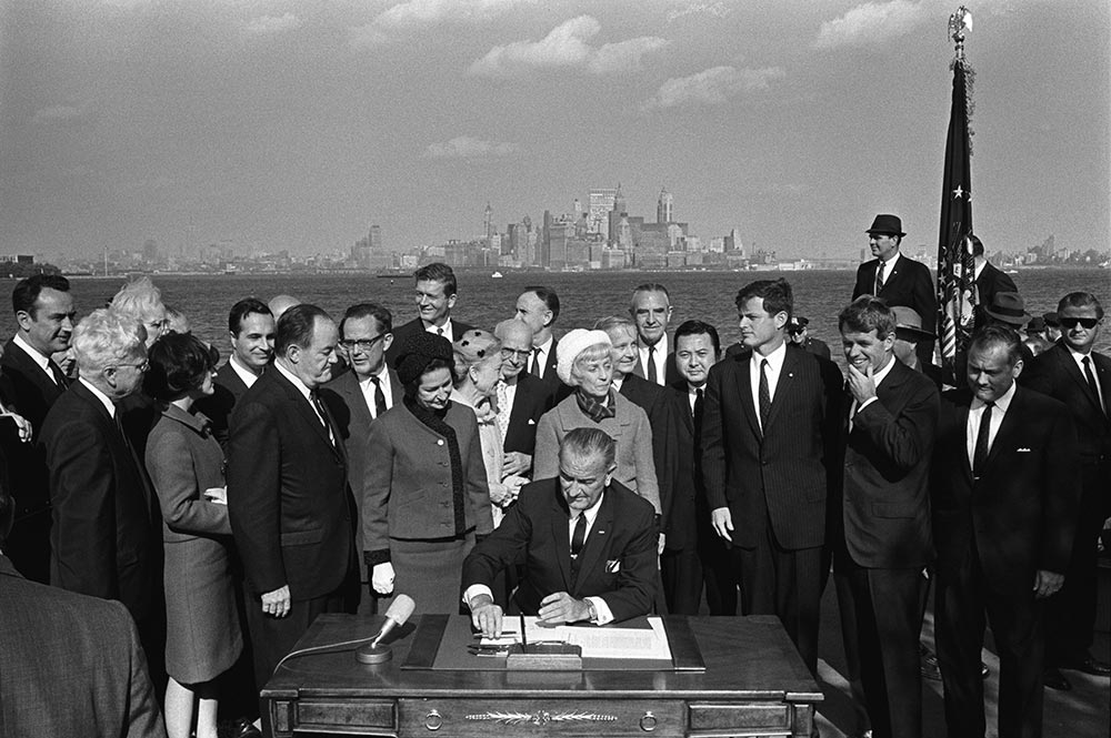 President Lyndon B. Johnson signs the Immigration Act as Vice President Hubert Humphrey, Lady Bird Johnson, Muriel Humphrey, Sen. Edward Kennedy, Sen. Robert F. Kennedy, and others look on, photograph by Yoichi Okamoto, October 3, 1965 (White House Photo Office / Lyndon Baines Johnson Presidential Library)