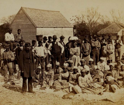 Group of emancipated enslaved people sitting in yard. With buildings visible in background.