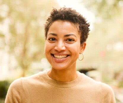 A headshot of a smiling Black woman with short hair wearing small gold hoop earrings and a tan sweater