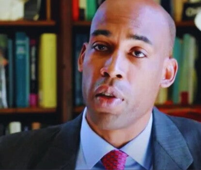 A headshot of a Black man speaking, wearing a black sport coat, light blue shirt, and patterned red tie, in front of a wall of books