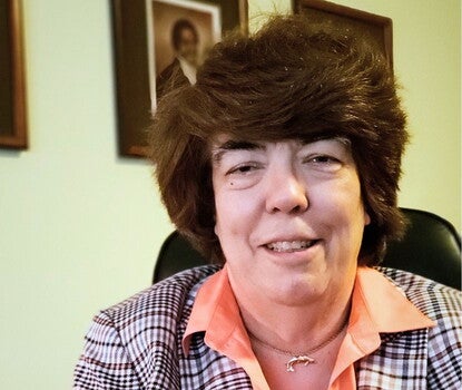 A headshot of a smiling White woman with short dark hair, wearing a plaid jacket and light orange shirt; to her right, an inset of the cover of the book "Between Slavery and Freedom."
