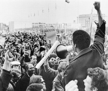  A Black man wearing a dark coat speaking into a bullhorn megaphone held in his left hand, while his right arm is outstretched with a clenched fist. A crowd looks on and raises their right fists in the air. There is a sign reading "Viva La Huelga" in the left midground.