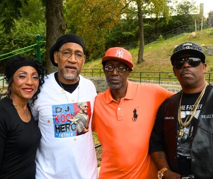 A color photograph of four people in a park: a Black woman wearing a black shirt and headband; a Black man wearing a white t-shirt, chain with large star, black undershirt, glasses, and backward black baseball cap; a Black man wearing a red polo shirt, black plastic-frame glasses, and a red New York Yankees cap; and a Black man with a black vest and t-shirt, two chains, sunglasses, and a black baseball cap.