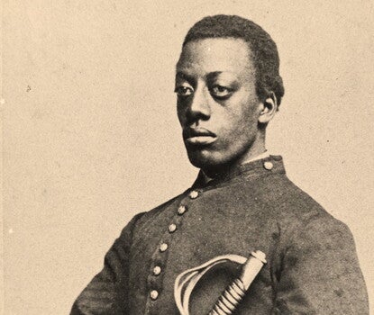 A black-and-white photograph of a serious young Black man in a military uniform (dark coat, white gloves, light-colored pants with a dark racing stripe) holding a sheathed sword