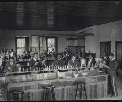A photograph of a chemistry laboratory, with three lab benches in the foreground, midground, and background. Black men in white shirts, dark coats, and dark ties are working at each bench. In the right foreground, framed by a doorway behind him, is George Washington Carver, a mustachioed Black man wearing a dark coat and tie, looking down at another chemist's work.