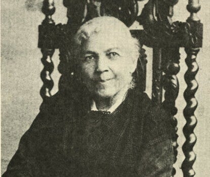 A smiling white-haired Black woman in a dark dress sitting with her hands clasped in her lap in an elaborately carved high-backed wooden chair