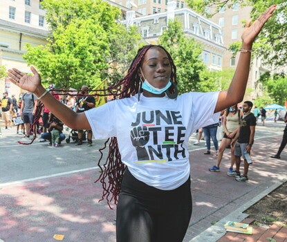  A color photograph of a young Black woman with arms outstretched, a surgical mask under her chin, wearing a white shirt bearing the emblem of black fist and the word Juneteenth with trees, tall buildings, and groups of people behind her