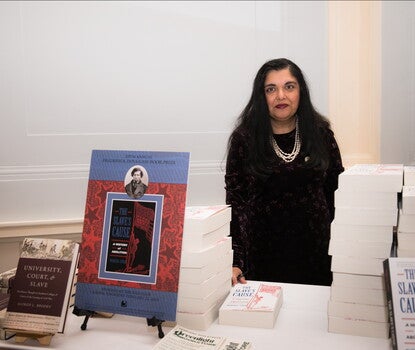 A South Asian woman wearing a black dress, black sweater, and white necklace, behind a table with paperback copies of the book "The Slave's Cause: A History of Abolition"