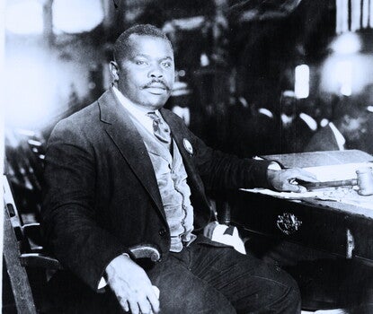 A black-and-white photograph of Marcus Garvey, a Black man wearing a dark suit, light-colored vest, white shirt, and dark tie, sitting in a wooden chair next to a desk filled with papers, holding a handkerchief in his right hand and a gavel in his left.
