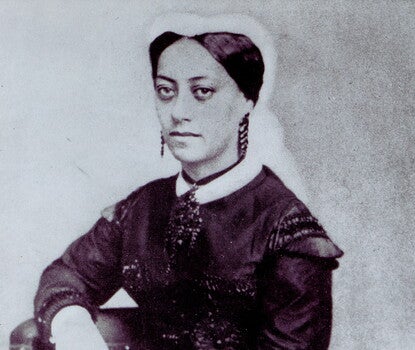 A black-and-white photograph of a serious-looking, dark-haired Black woman wearing a dark coat, dress with a white collar, earrings, and a white veil on the back of her head