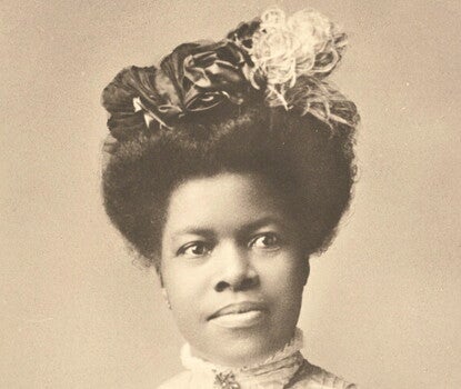A black and white photograph of Nannie Helen Burroughs, a young Black woman, shown seated from the waist up, wearing a dark hat with a white feather and a white blouse