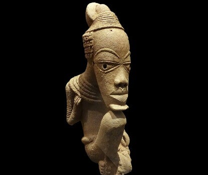 A stylized terracotta sculpture of a seated man with an oversized and elongated head relative to their body, chin resting on the figure's knees. Damage to the figure's shins.