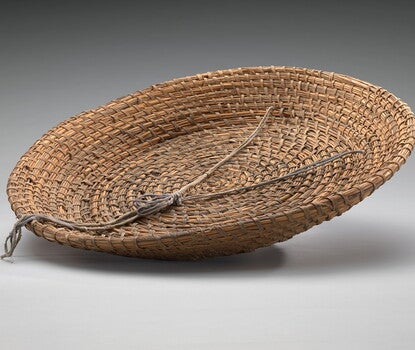 A large shallow, round tray shaped basket made of reeds, rush and palmetto coiled and woven together. A faded purple string is woven through the basket on one side and knotted inside the basket.