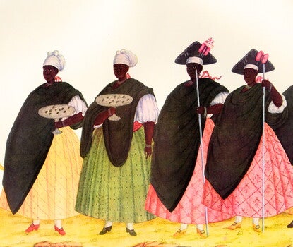 A group of five Black women wearing white and black headdresses, black cloaks, white blouses, brightly colored skirts, and high-heeled shoes, follows a small boy wearing colorful clothing adorned by feathers, holding a piece of wood and a small ax. 