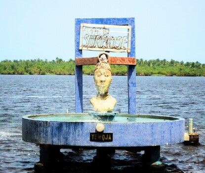 A jet of water in a tiled water fountain labeled 'Yemoja" [also known as Yemayá] in a body of water, topped with a white bust of a woman and, above the bust, a dark neon sign reading "The Surprise."