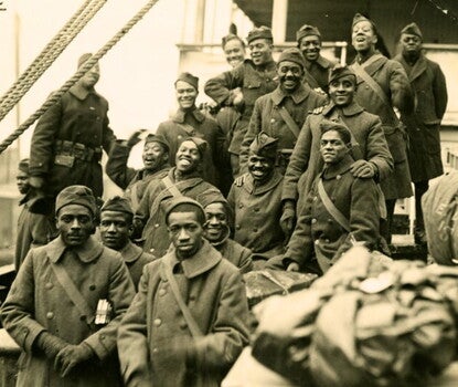 A black-and-white photograph of several Black men in army uniforms (dark double-breasted coats, dark gloves, dark hats) standing aboard a ship, rigging visible in the left midground and cargo in the right foreground and midground.