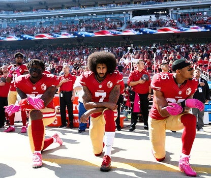 Three of the San Francisco 49ers football team kneeling (right knee on ground, arms balanced on left knee) during the pregame performance of the national anthem, as their teammates, coaches, and staff stand, some with hats or hands on their left breast, behind them. The three Black men kneeling are wearing the 49ers uniform of red jerseys, gold pants (with red-white-red stripes along the leg), and either red or white socks.