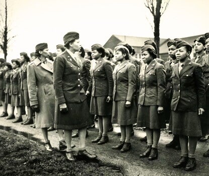 Two Black women in military uniform (dark jackets, light shirts and ties, dark skirts and boots) looking on at two rows of other Black women in military uniform standing at attention.