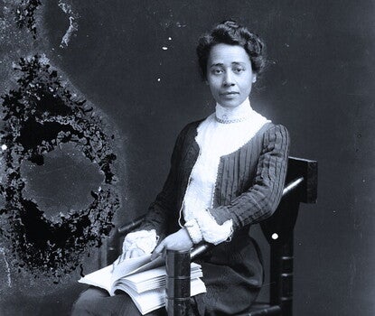 Anna Julia Cooper, a Black woman, wearing a black dress with a white center placket, sitting while holding a book.