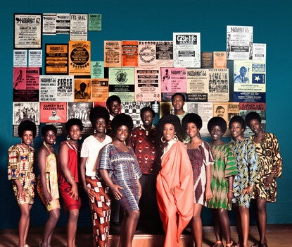 A color photograph of a row of ten female Grandassa models with natural, afro-style hair wearing dresses made from brightly colored African-inspired motifs. Three men standing on a small podium behind the models are wearing patterned dashiki shirts. The dark blue wall behind them is covered with posters advertising Black theater performances, shows, and celebrations.