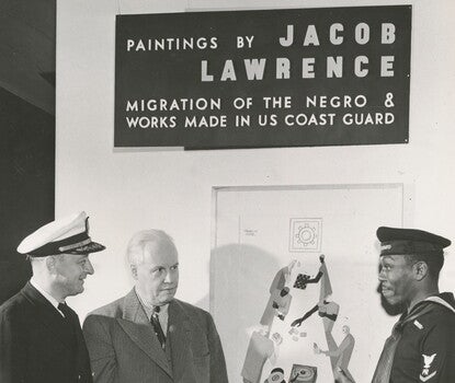 Black-and-white photograph of an art gallery, with three men standing in the gallery: a white man in his Coast Guard captain uniform of dark double-breasted coat, white shirt, dark tie, and white cap with a dark brim); at center, a white man in a dark suit, light colored shirt, and dark tie; and at right, a Black man in his Coast Guard sailor uniform of dark shirt (with gold bands on the sleeves) and pants, light-colored scarf, and dark beret.