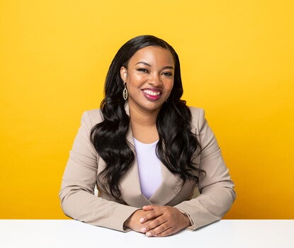 Ayesha Rascoe, a Black woman, is smiling, wearing a tan suit with a white scoop neck shirt, teardrop earrings, and bright pink lipstick. The background is a bright yellow backdrop.