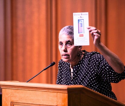 Barbara Smith, a Black woman, wearing a polka dot (white dots on black fabric) shirt and speaking into a lectern microphone. In her left hand, she is holding a pamphlet entitled "United States of All."