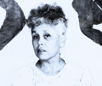 Betye Saar, a Black woman, wearing a white shirt with black streaks, her arms crossed. Prints of her art are in the background behind her.