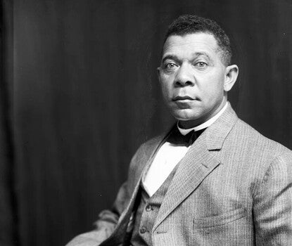 Booker T. Washington, a Black man, wearing a light colored jacket, vest, and pants, white shirt, and black bow tie.