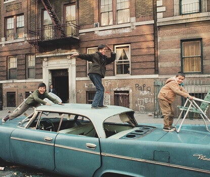Three boys playing atop the roof of a blue car,  in front of an apartment building with graffiti written on the ground floor.
