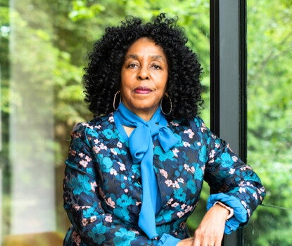 A photograph of a Black woman with black curly hair and large hoop earrings, wearing a blue flowered blouse and a cerulean ascot posed in front of a window.