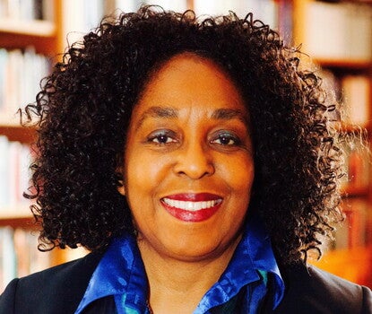 A Black woman with black curly hair and red lipstick smiles at the camera. She wears a collared blue patterned shirt, and a suit jacket. 
