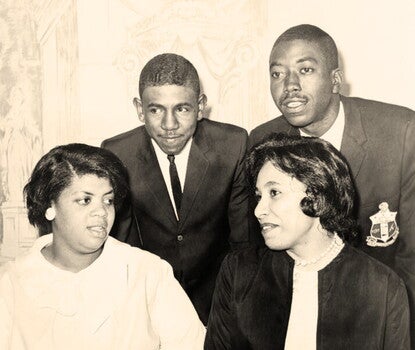 From left to right, a seated Black girl wearing a white coat and earrings; a standing Black boy wearing a dark jacket, white shirt, and thin dark tie; a seated Black woman, wearing a dark jacket over a light colored blouse and a pearl necklace; and a standing Black man, wearing a blazer emblazoned with a crest.