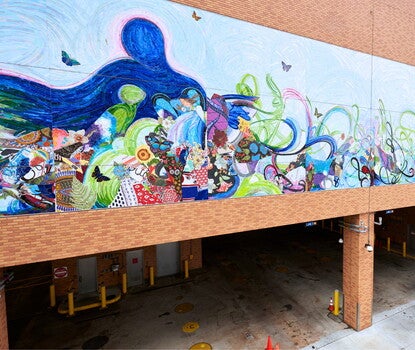 An abstract multicolored mural above a bus depot, with a blue and white bus pulling into the depot in the right foreground.