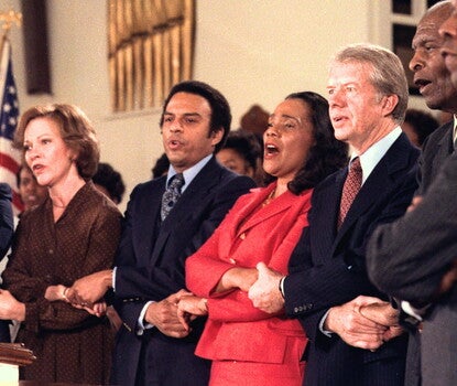 A group of five Black men and women hold hands and sing with a white man and woman inside a church. 