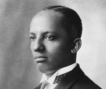 Carter G. Woodson, a Black man, wearing a dark jacket, white shirt (with a small collar), and polka-dotted tie (white dots on a dark fabric) with a tie bar or button.