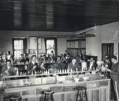 A photograph of a chemistry laboratory, with three lab benches in the foreground, midground, and background. Black men in white shirts, dark coats, and dark ties are working at each bench. In the right foreground, framed by a doorway behind him, is George Washington Carver, a mustachioed Black man wearing a dark coat and tie, looking down at another chemist's work.