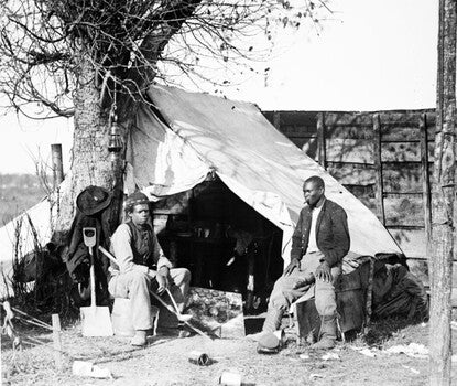Two African American men sitting in front of a tent, one with cigar and the other with a soup ladle.