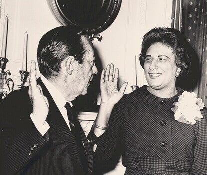 Black and white photo of a middle-aged Black woman with shoulder length hair and wearing a large white flower pin being sworn in as the borough president of Manhattan. 