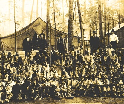 Several men (mostly Black men) in striped jumpsuits sitting and standing. Several of the individuals have dogs sitting with them. Standing behind the prisoners are five (5) men in civilian clothes. In the background is a large tent and a small wooden building among trees. White text in a lighter colored rectangle at the bottom front margin reads "A North Carolina Convict Camp."