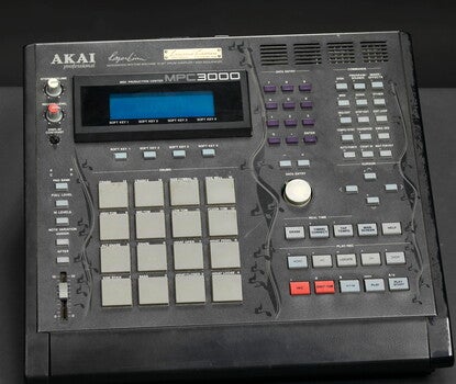 Photograph of a multi-buttoned drum machine and sampler. The machine consists of a black plastic box with a small screen in the upper left corner, multiple key pads, and a vertical design of black musical notes along the left, center and right of the box.