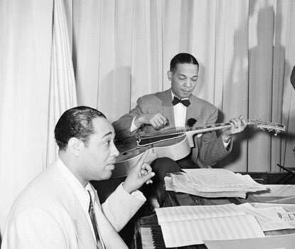 Duke Ellington, a Black man wearing a light colored jacket, white shirt, and dark tie, playing the piano with his right hand and conducting with his left as a Black man wearing a tuxedo plays guitar in the background.