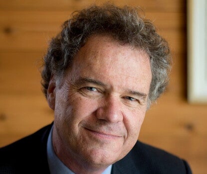 Photograph of a man with graying, curly hair, smiling at the camera in front of a wooden wall. He wears a dark suit jacket, a blue collared shirt, and a red and blue paisley tie. 