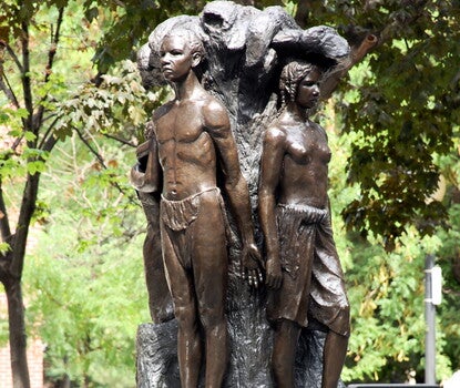 A photograph of a bronze cast sculpture titled "Emancipation." The sculpture depicts male and female figures emerging from the tree of knowledge at the center.