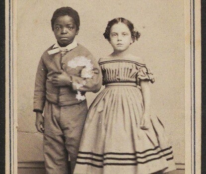  A photograph of a young dark-skinned boy wearing a jacket, slacks, arm-in-arm with a light-skinned girl wearing a light-colored dress